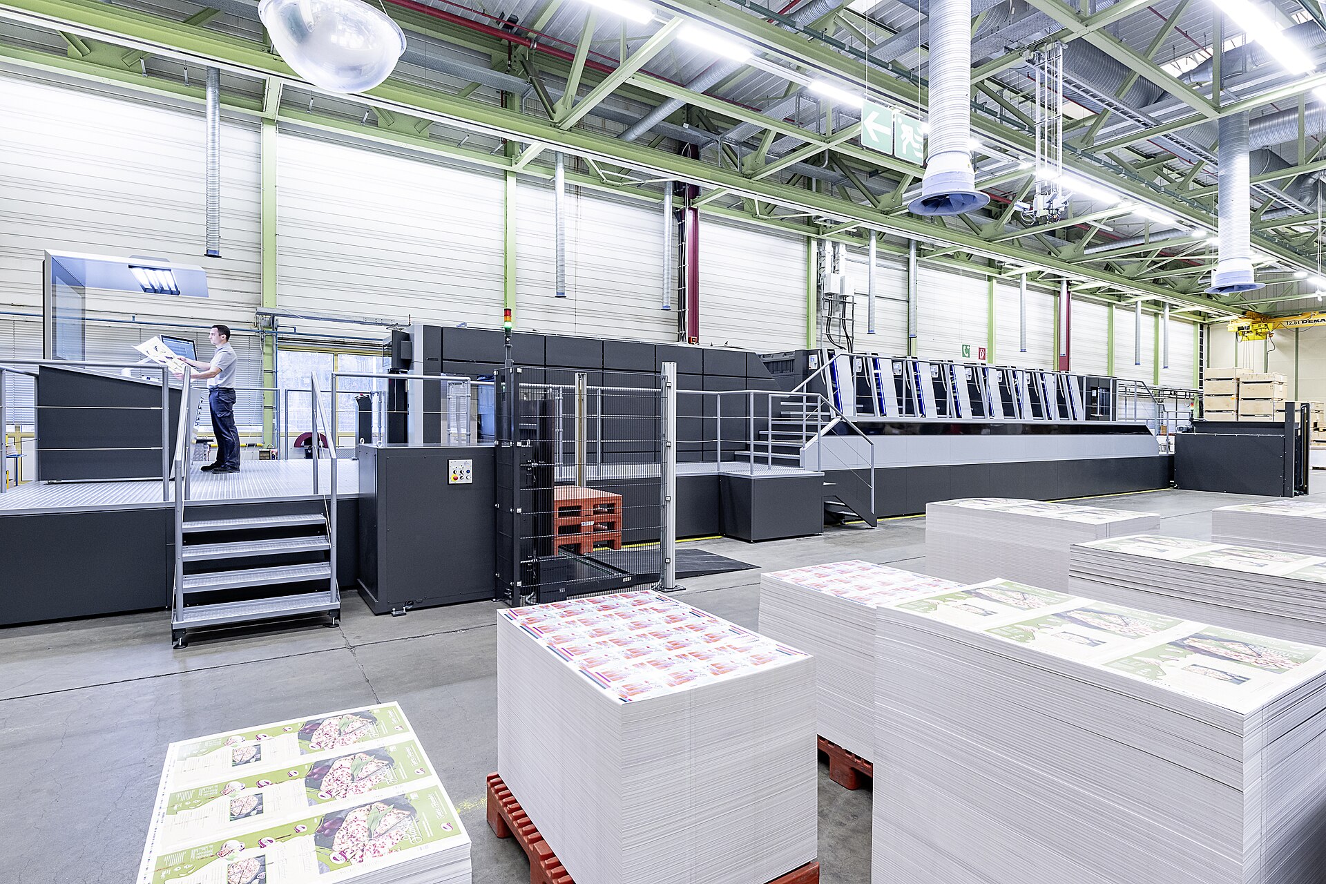 Press hall with an operator checking a printed sheet beside stacks of printed stock